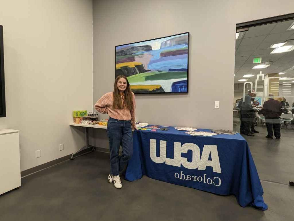 An intern stands in front of a table with a blue ACLU of Colorado tablecloth and an assortment of printed materials